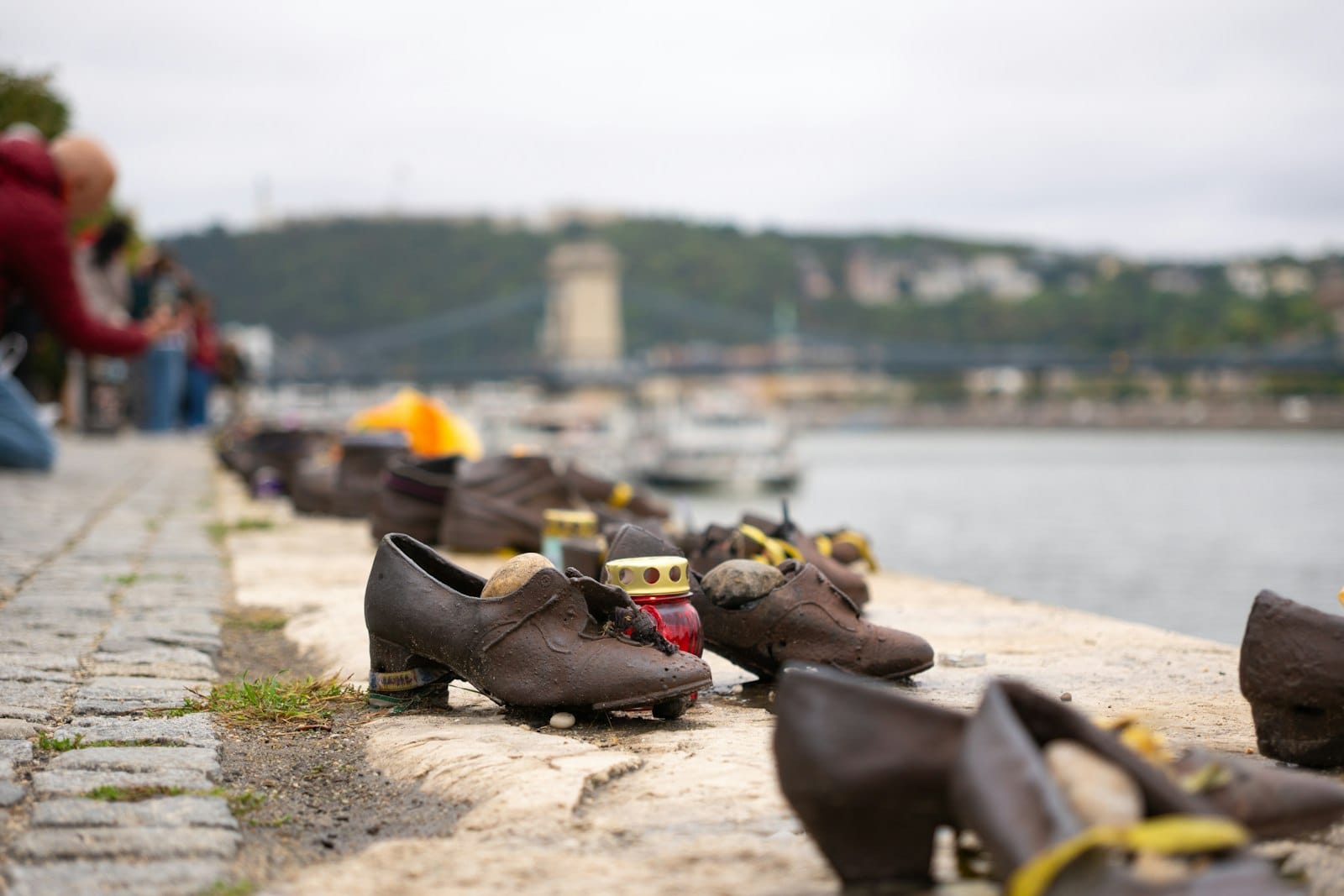 Malapit na tingin sa mga bronze na sapatos na nakahanay sa tabi ng ilog na memorial sa Budapest na may tulay at tanawin ng lungsod sa likod.