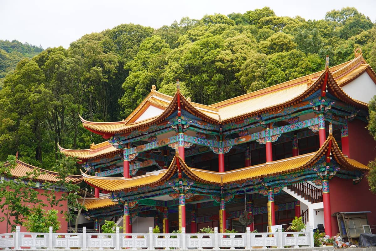 Zweistöckiges traditionelles chinesisches Tempelgebäude mit verzierten Balken, bunten Dekorationen, roten Säulen und Wald im Hintergrund.