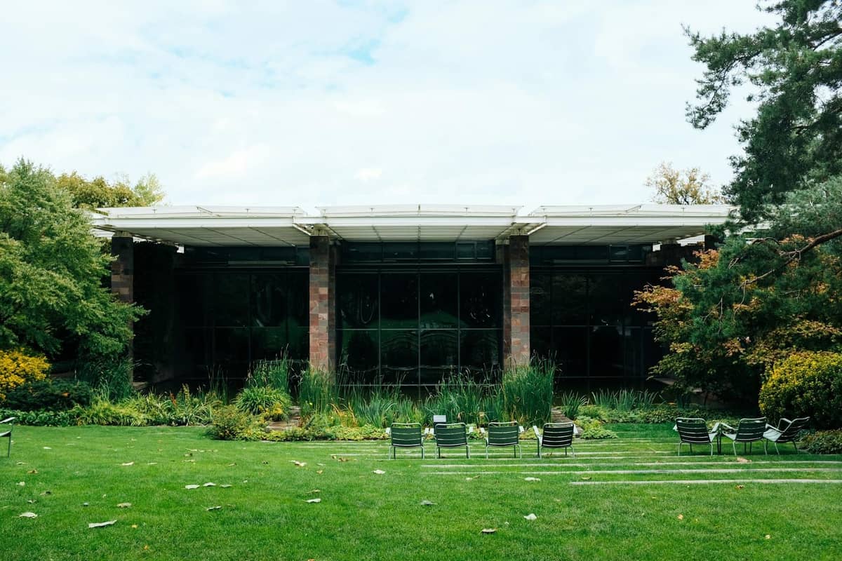 Padiglione modernista in vetro immerso in un prato verde e un giardino paesaggistico, con file di sedie all'aperto e alberi su entrambi i lati.