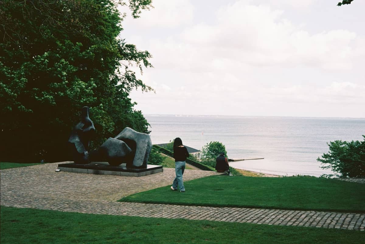 Park rzeźb z figurą nowoczesnej sztuki, ścieżką, bujnym zielonym trawnikiem i widokiem na morze, spacerujący ludzie w pobliżu Louisiana Museum.