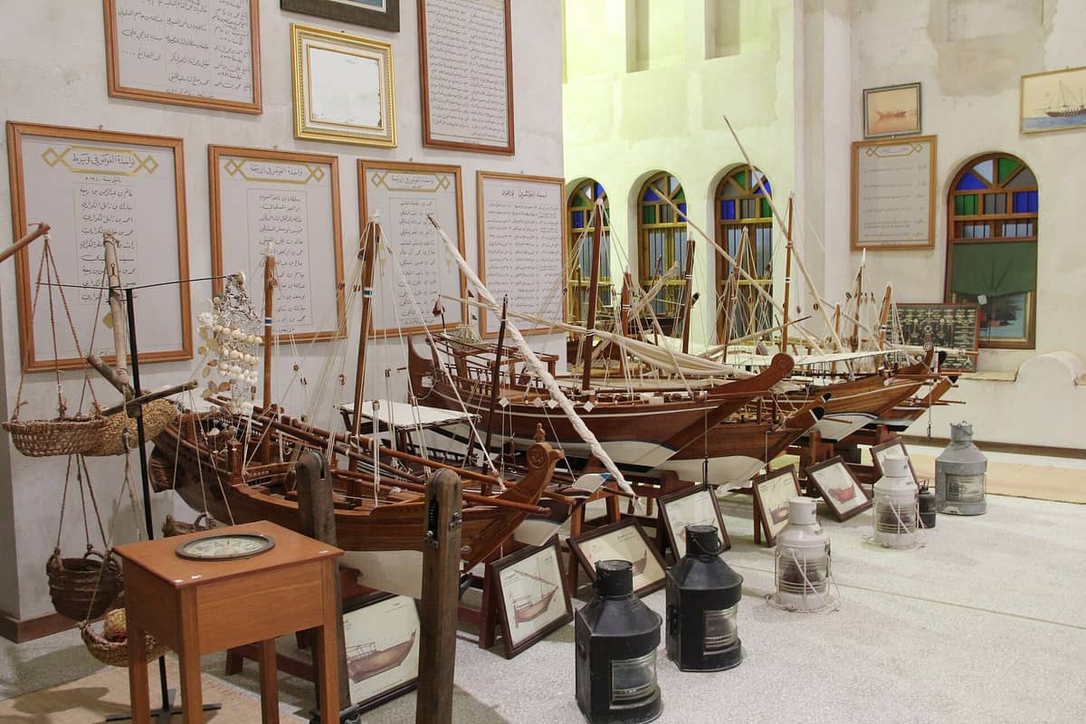 Ausstellung traditioneller hölzerner Dhow-Bootsmodelle und Perlentauch-Artefakte in einem Kulturerbemuseum in Doha, mit arabischem Text und bunten Bogenfenstern.