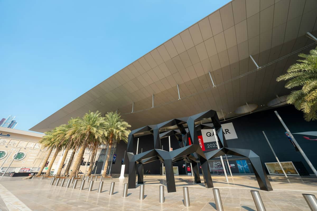 Weitwinkelansicht des Außenbereichs des 3-2-1 Qatar Olympic and Sports Museum mit Palmen, moderner Kunstskulptur und Museumseingang unter einem markanten Dach.