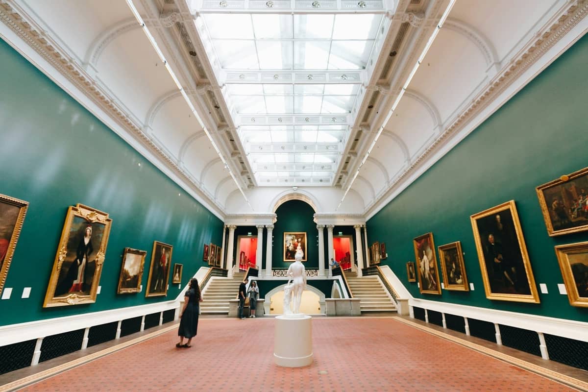Vista interior de una gran galería con paredes verdes, gran claraboya, pinturas clásicas y visitantes admirando el arte.