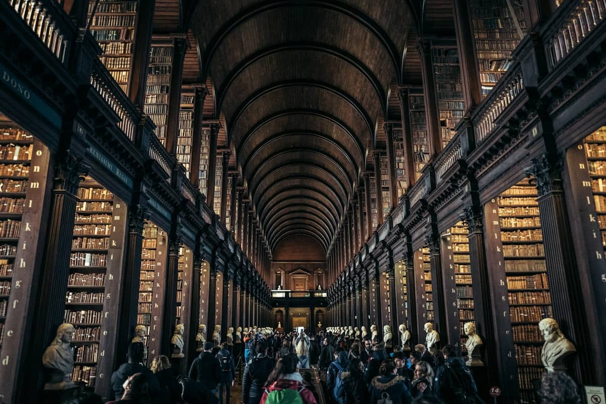 La Long Room en la Biblioteca del Trinity College, Dublín, con altas estanterías, techo de madera abovedado, bustos de mármol y visitantes caminando por ella.