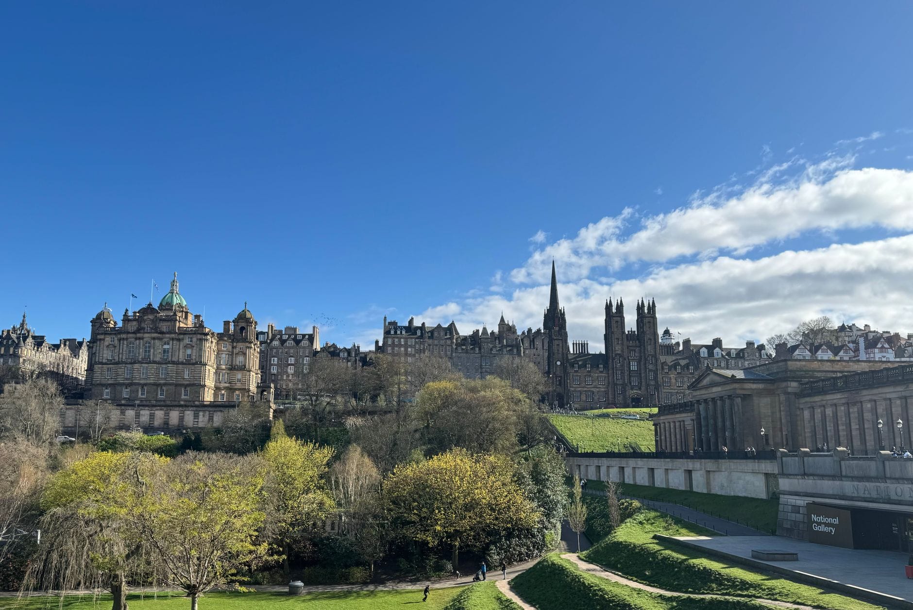 Panoramablick auf das Stadtzentrum von Edinburgh mit historischen Gebäuden und Türmen, mit dem Gebäude der National Gallery und üppigen Gärten im Vordergrund.