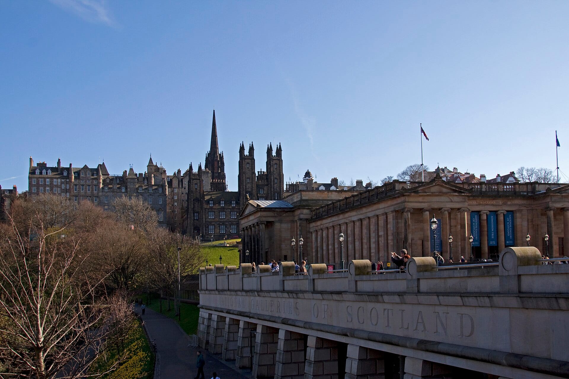 Blick auf das Äußere der Scottish National Gallery mit Menschenmassen davor und der Skyline der Altstadt von Edinburgh im Hintergrund an einem sonnigen Tag.