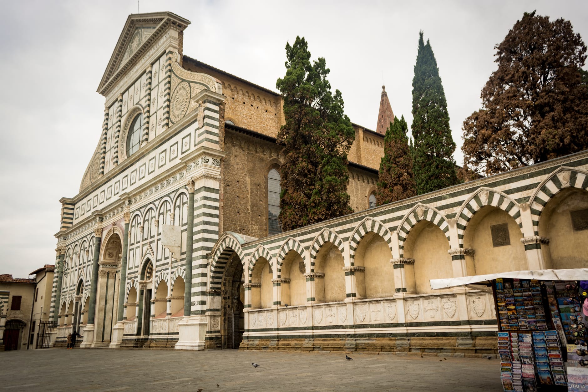 มุมมองแบบไวด์แองเกิลของมหาวิหาร Santa Maria Novella ในเมืองฟลอเรนซ์ แสดงให้เห็นด้านหน้าอาคารที่ประดับประดาอย่างวิจิตรและระเบียงโค้งพร้อมต้นไม้ในพื้นหลัง