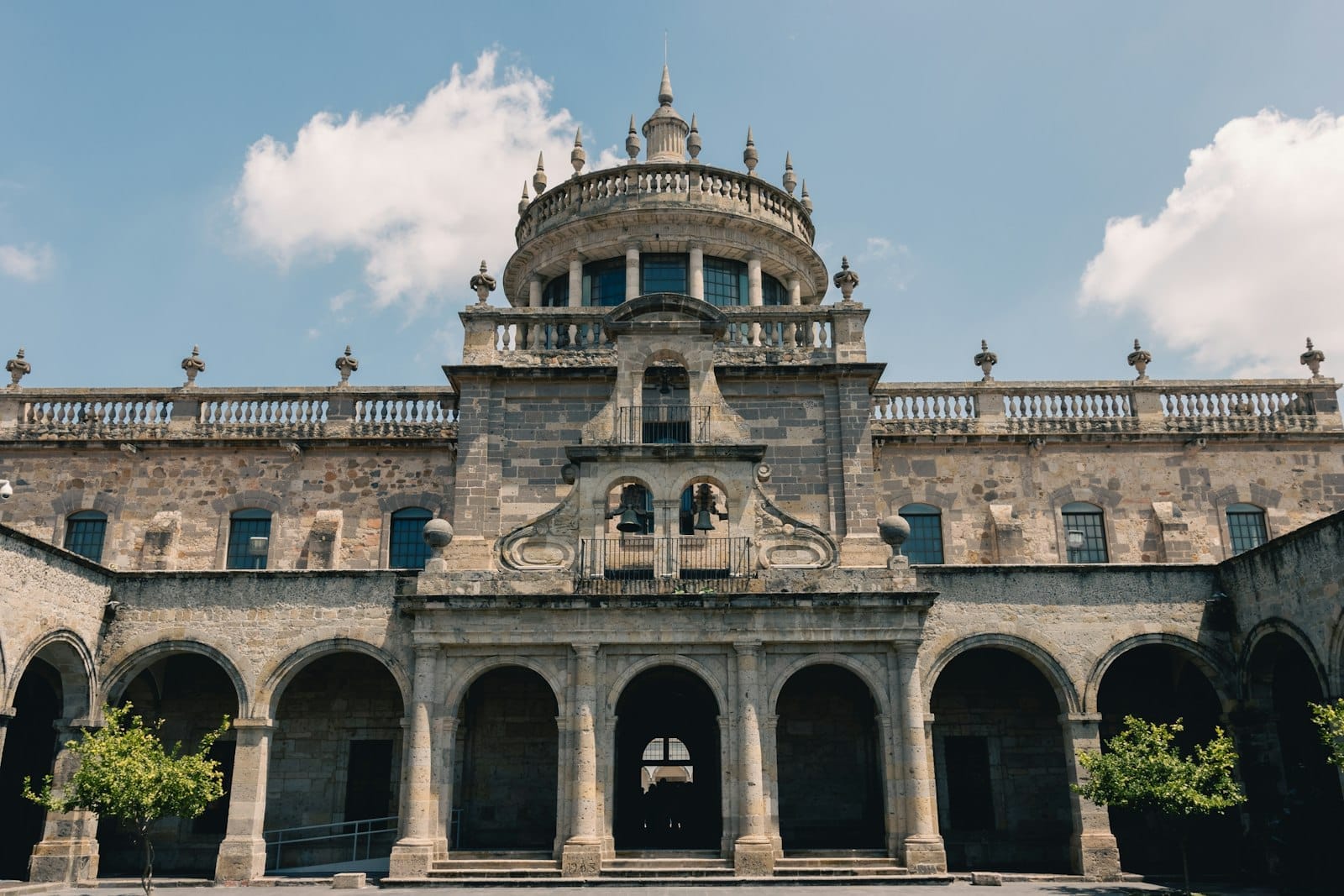 Vista frontal do Hospício Cabañas em Guadalajara em um dia ensolarado, com sua arquitetura histórica simétrica e entrada em arco sob um céu azul.