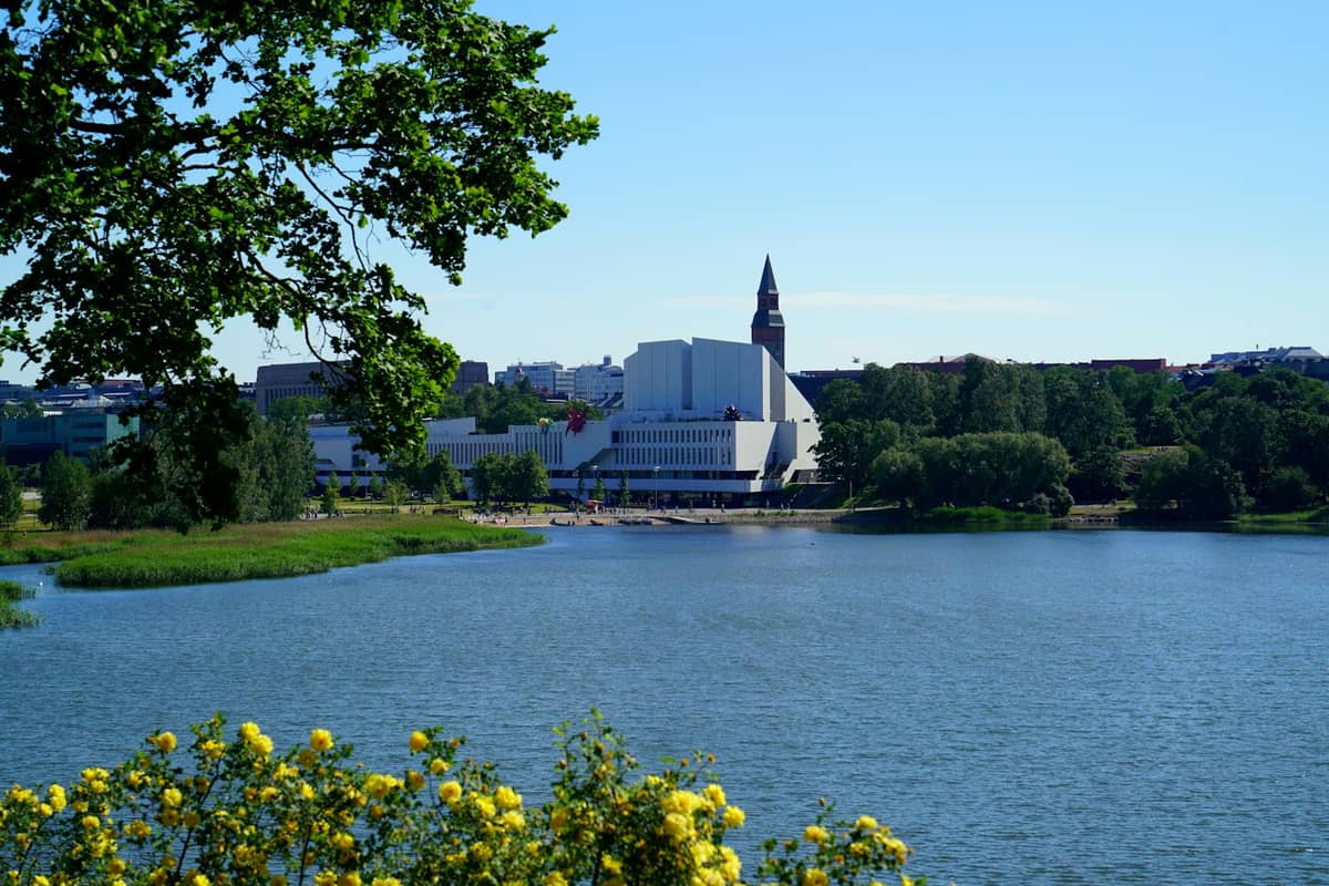 Vista de um lago para o Finlandia Hall e a torre do Museu Nacional em Helsínquia num dia ensolarado, com árvores e flores em primeiro plano.