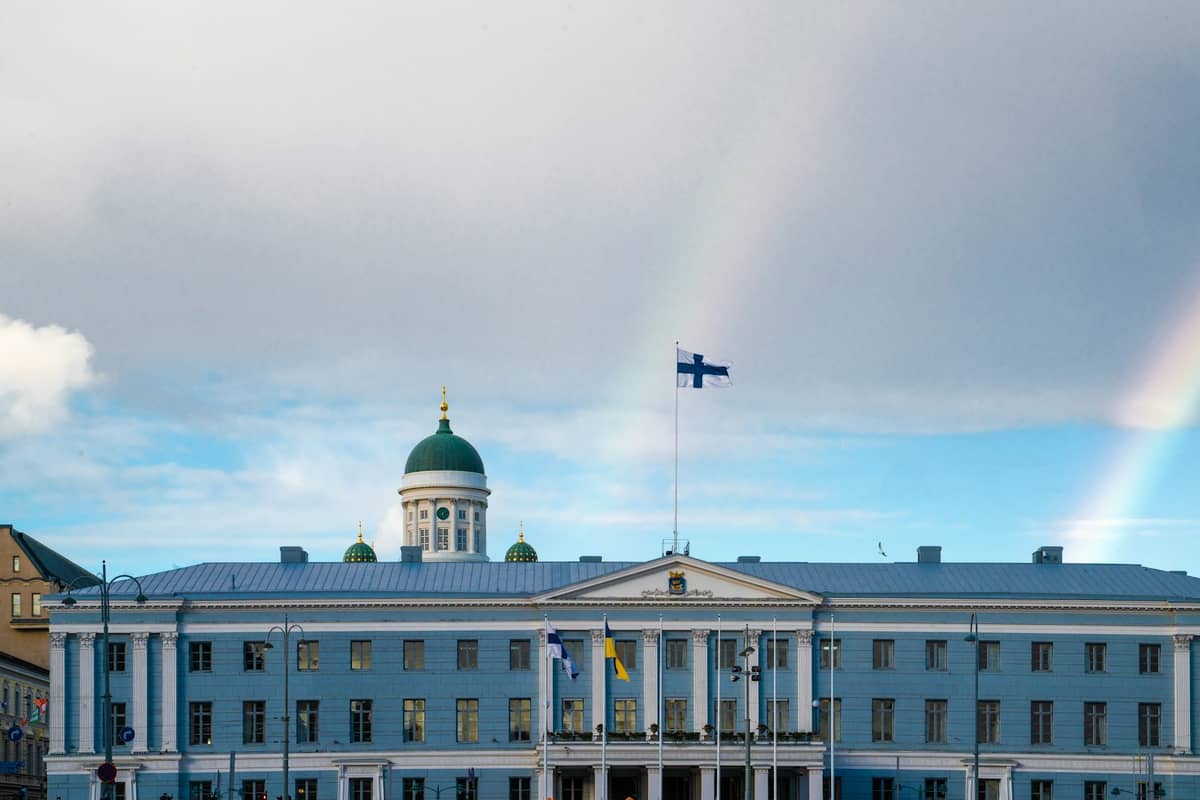 Um imponente edifício neoclássico com bandeiras finlandesas e suecas, com as cúpulas da Catedral de Helsinki visíveis e um arco-íris no céu nublado.