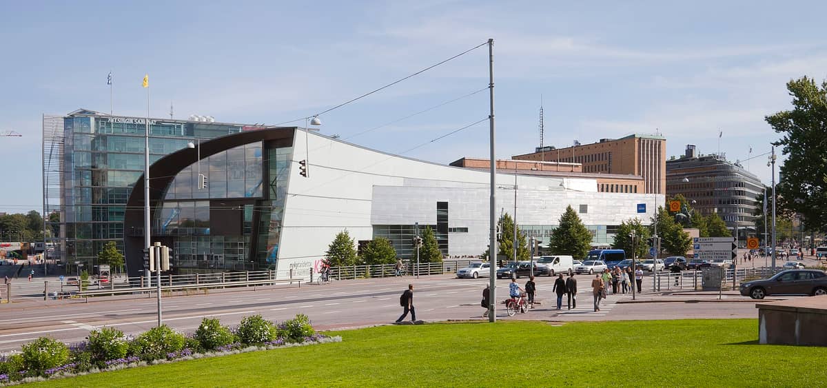 Vista ampla do edifício moderno e curvo do Kiasma, Museu de Arte Contemporânea, com pessoas atravessando a rua e prédios de Helsinki ao fundo.