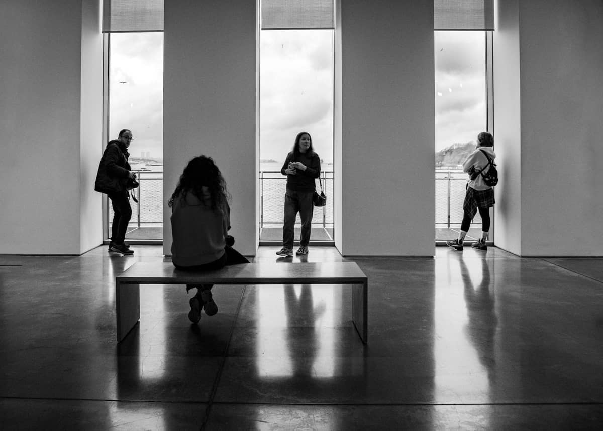Foto en blanco y negro de personas de pie y sentadas dentro de una galería de arte moderno con grandes ventanales y vistas al Bósforo.