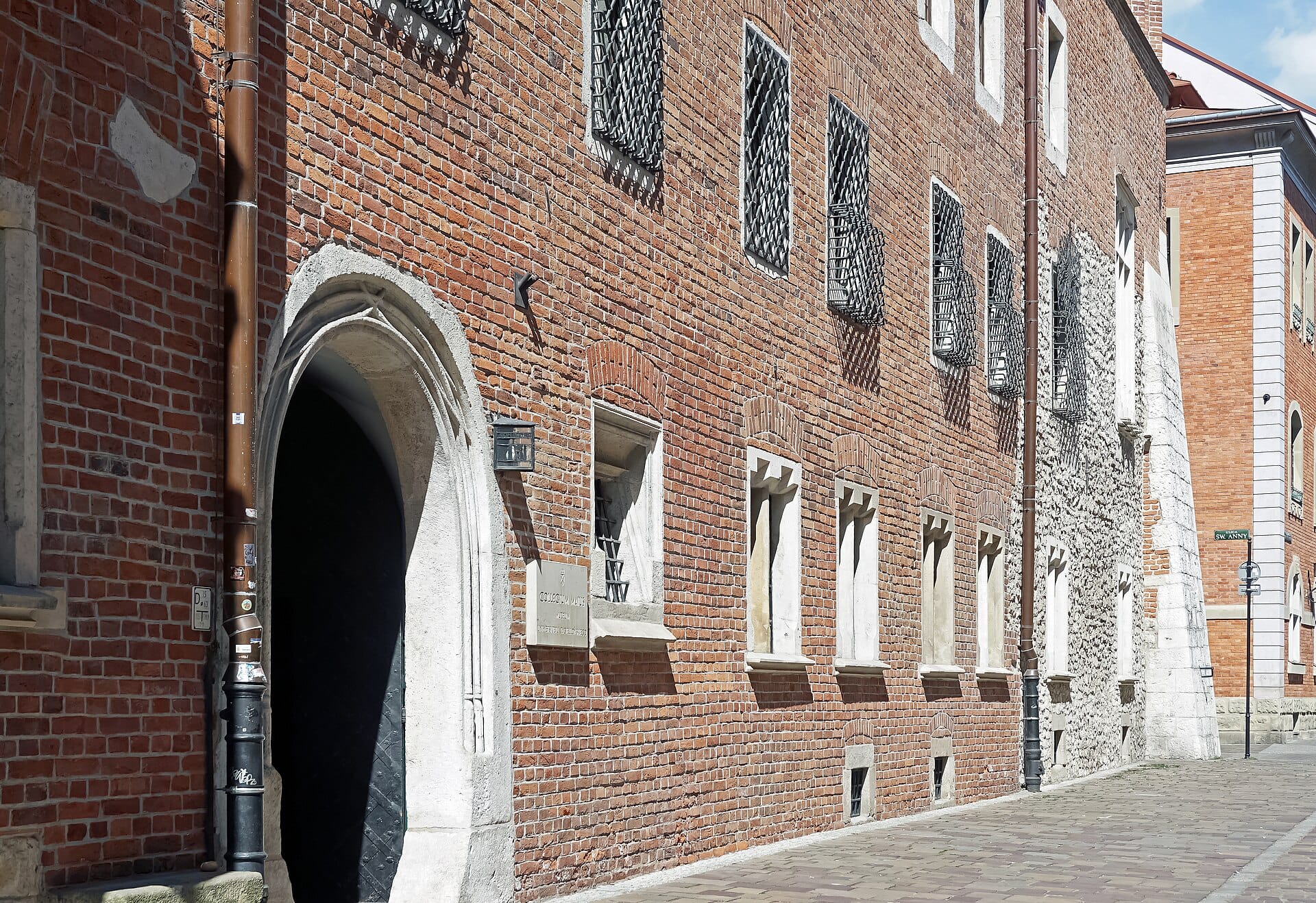 ด้านหน้าของ Collegium Maius เมือง Kraków พร้อมประตูหินโค้ง กำแพงอิฐสีแดง และหน้าต่างมีลูกกรงตามถนนที่เต็มไปด้วยแสงแดด