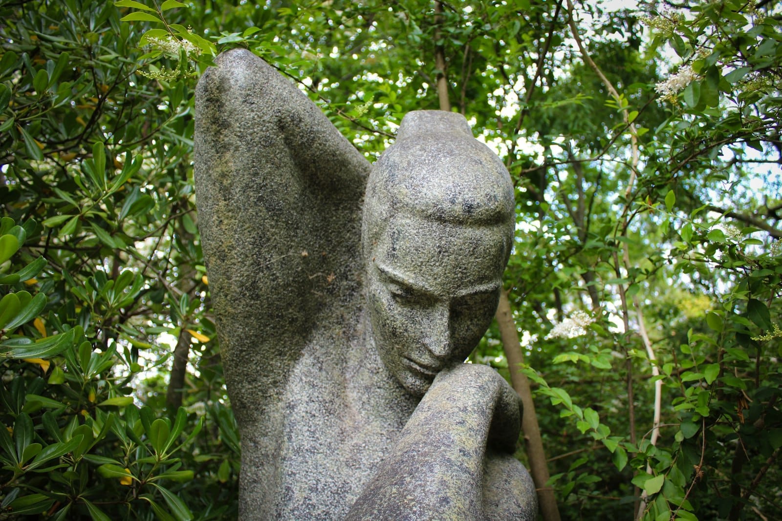 Gros plan d'une sculpture en pierre représentant une figure humaine, entourée de feuillage vert dans un jardin extérieur.