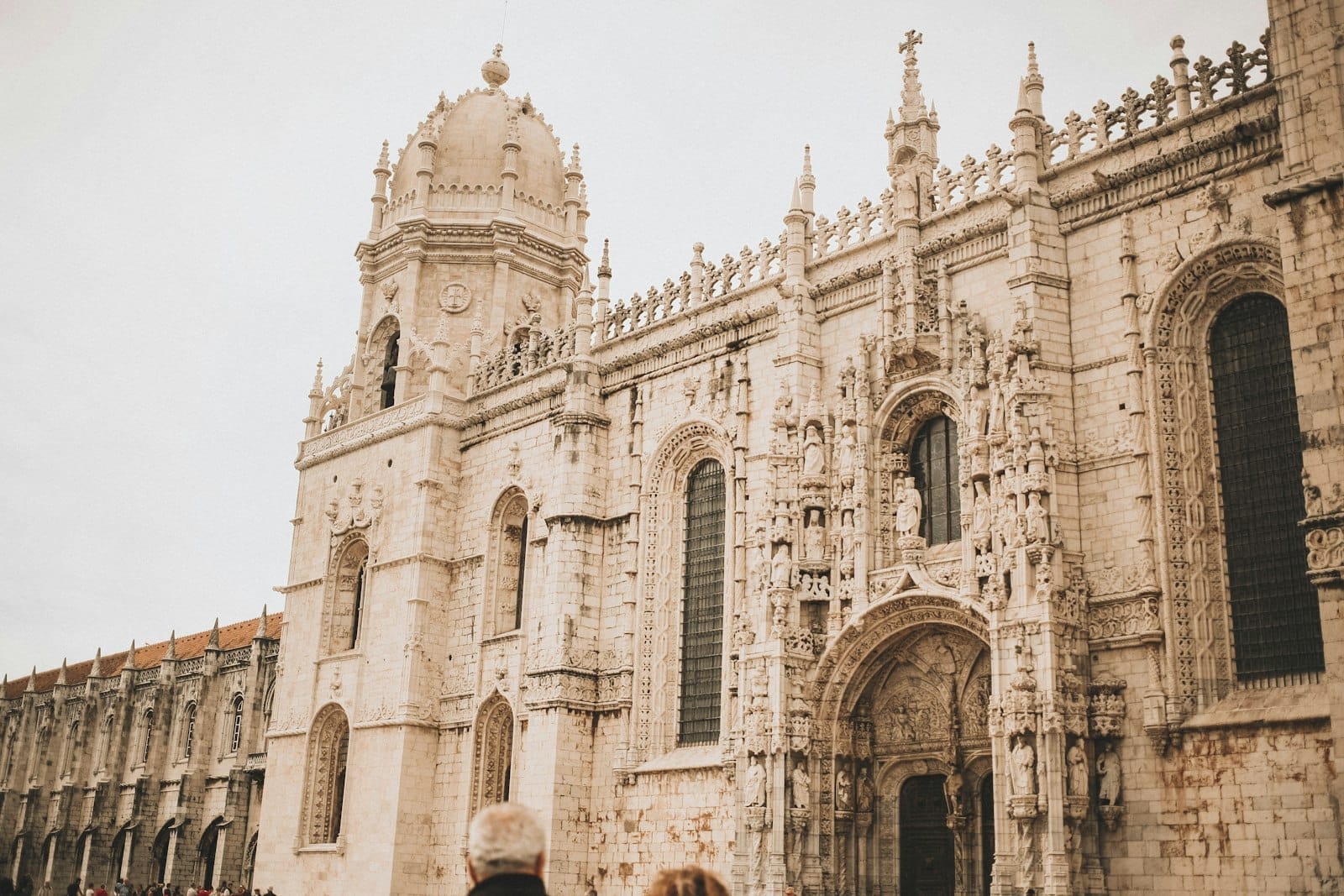 Façade en pierre détaillée et tour du Monastère des Jerónimos, avec une porte cintrée et des visiteurs à l'entrée sous un ciel nuageux.