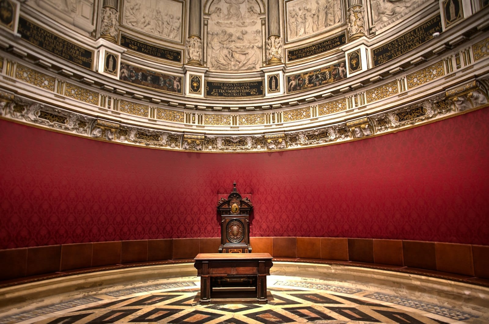 Salle du trône circulaire somptueuse en rouge et or avec un plafond ornemental et un unique fauteuil cérémoniel au centre.
