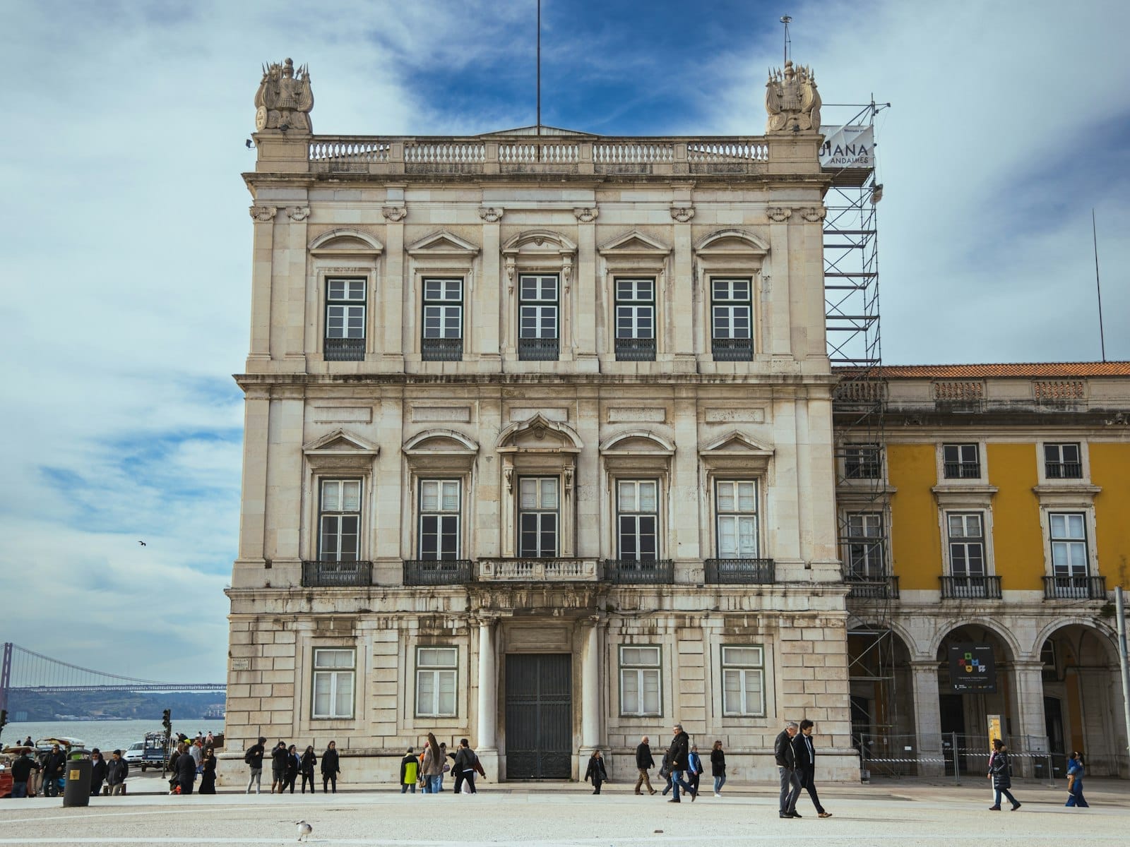 Bâtiment néoclassique orné sur la Praça do Comércio de Lisbonne, des personnes marchant devant, ciel bleu et vue partielle d'un bâtiment jaune à côté.