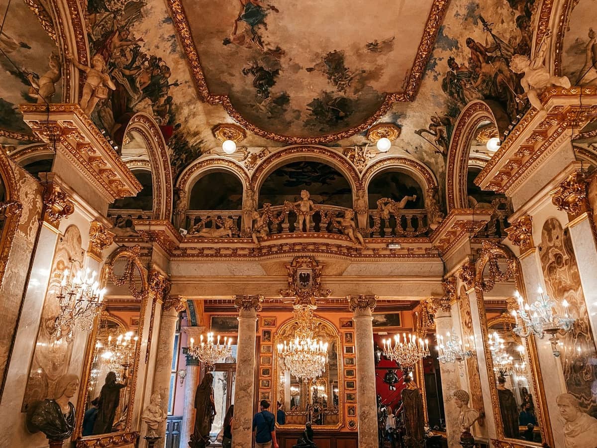 Interno di palazzo riccamente decorato con lampadari, finiture dorate ornate, soffitto dipinto e specchi, che mostra una grandiosità adatta a un museo di collezione privata.