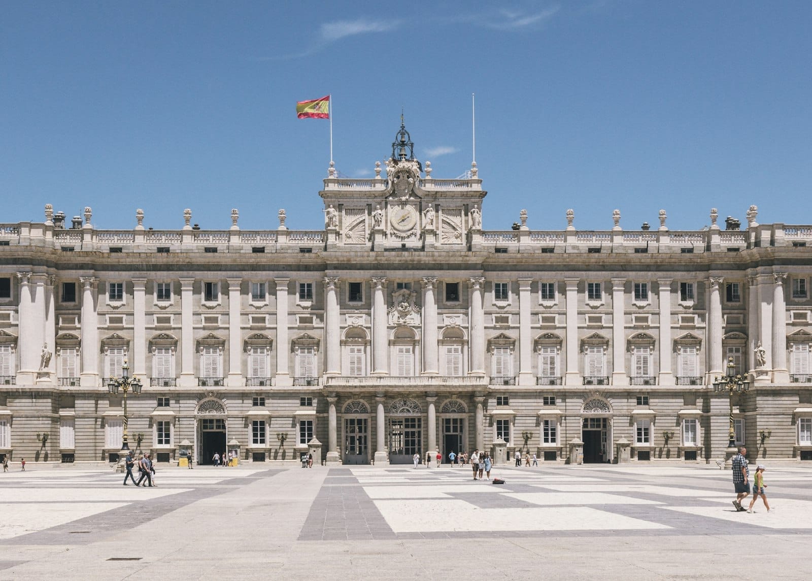 Vista ampla da fachada monumental do Palácio Real de Madri, com a bandeira espanhola hasteada e visitantes passeando em frente sob um céu azul límpido.