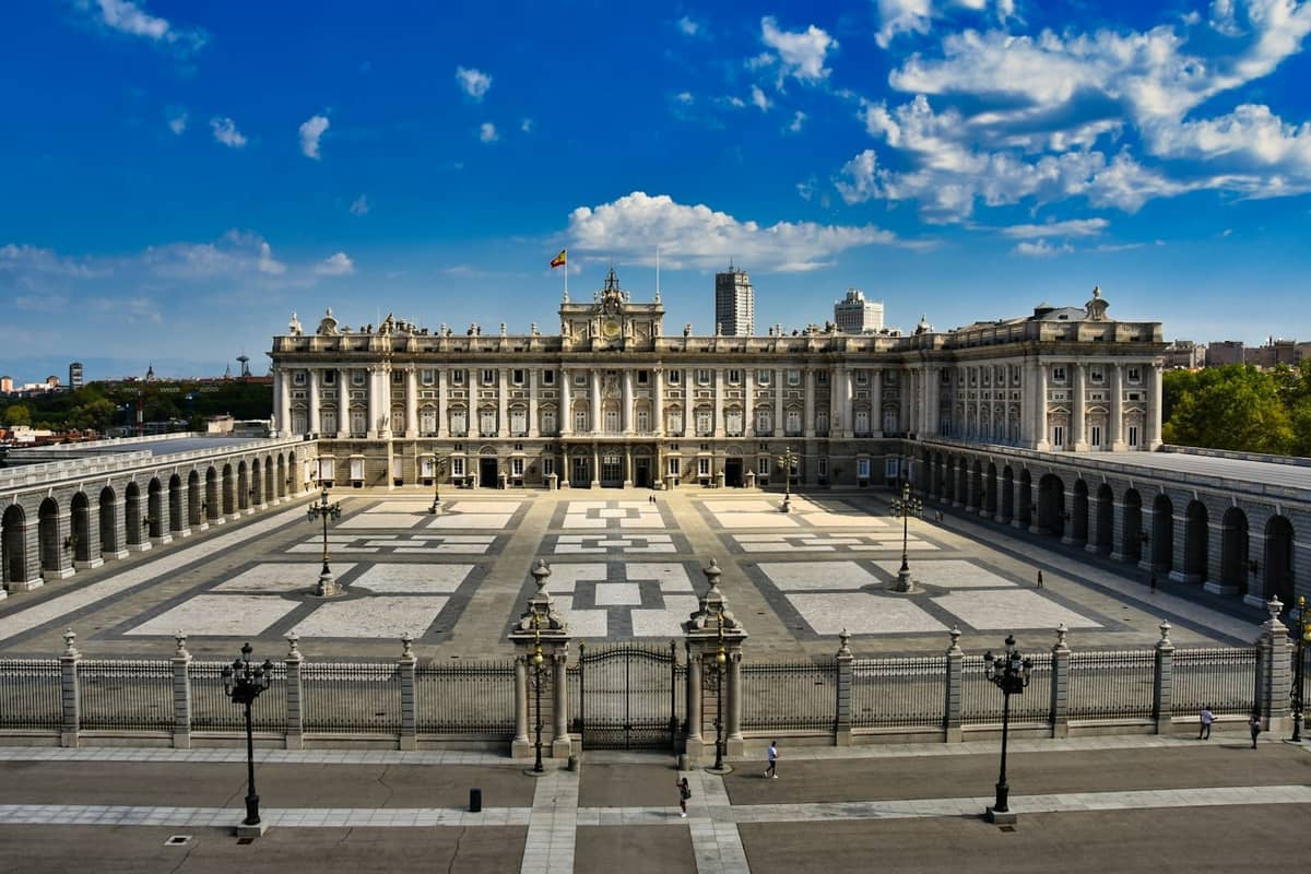 Ampia veduta del cortile del Palacio Real di Madrid sotto un cielo azzurro con nuvole sparse, che mostra l'imponente architettura storica e la vasta piazza.