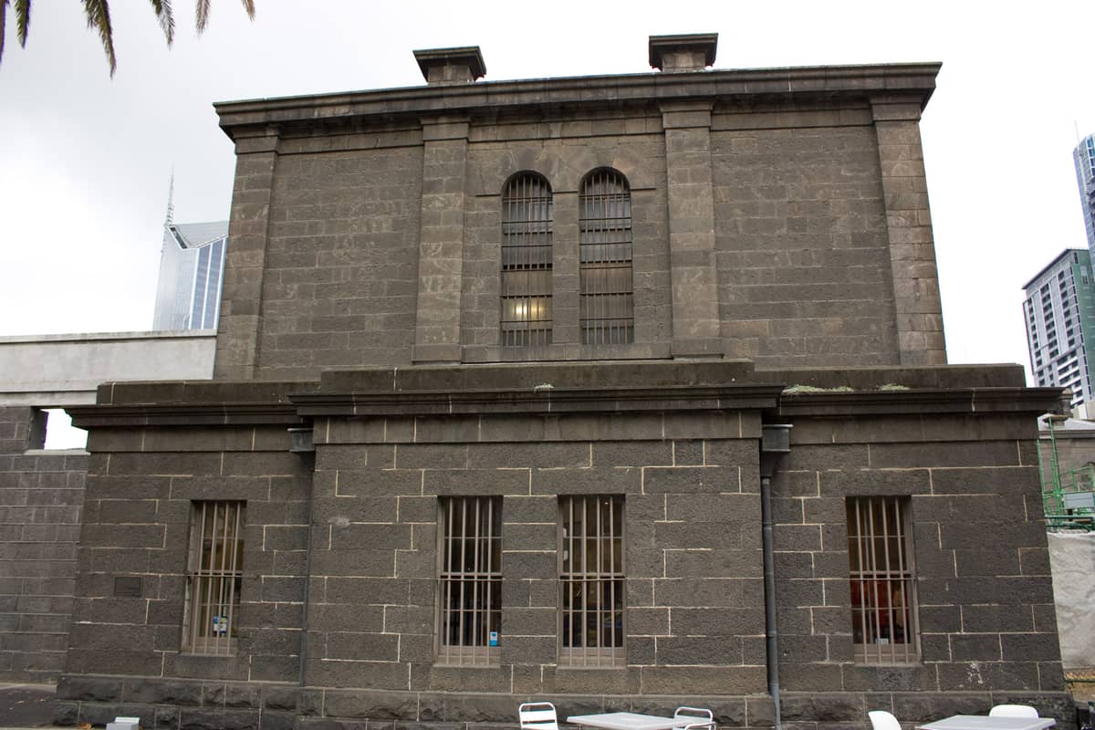 Fachada da Old Melbourne Gaol com grossas paredes de pedra azul e janelas gradeadas, um edifício histórico no centro de Melbourne.