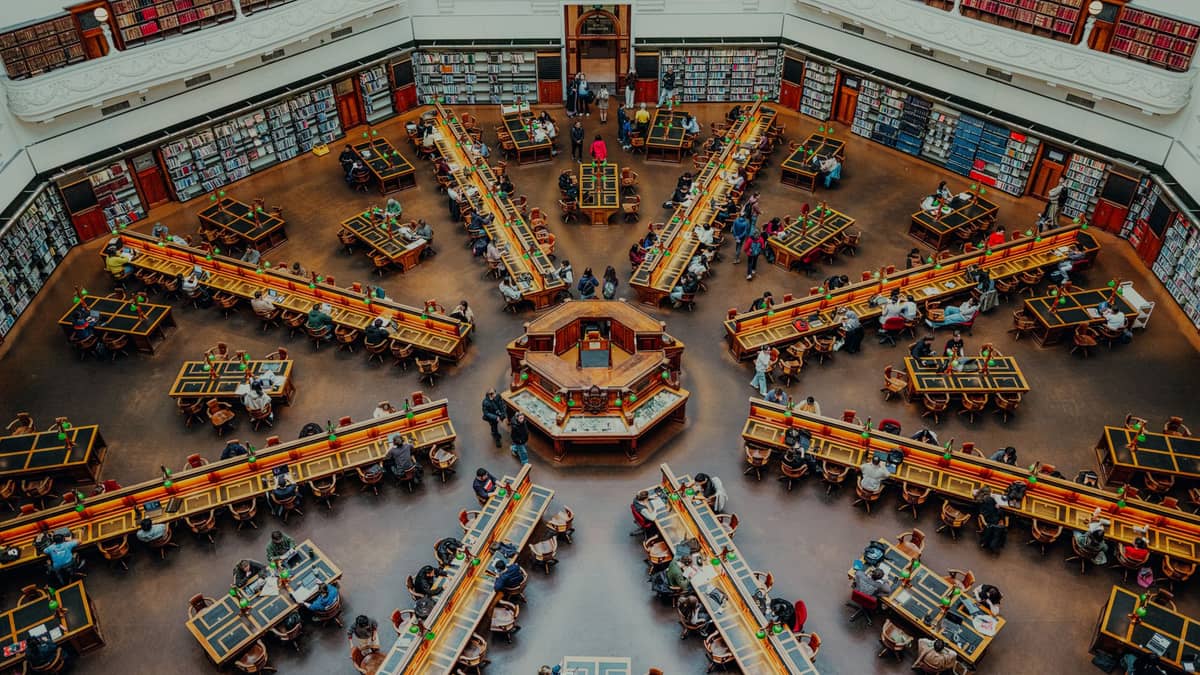 Vista ampla e aérea da Sala de Leitura La Trobe na State Library Victoria, com mesas octagonais, pessoas estudando e paredes repletas de livros.