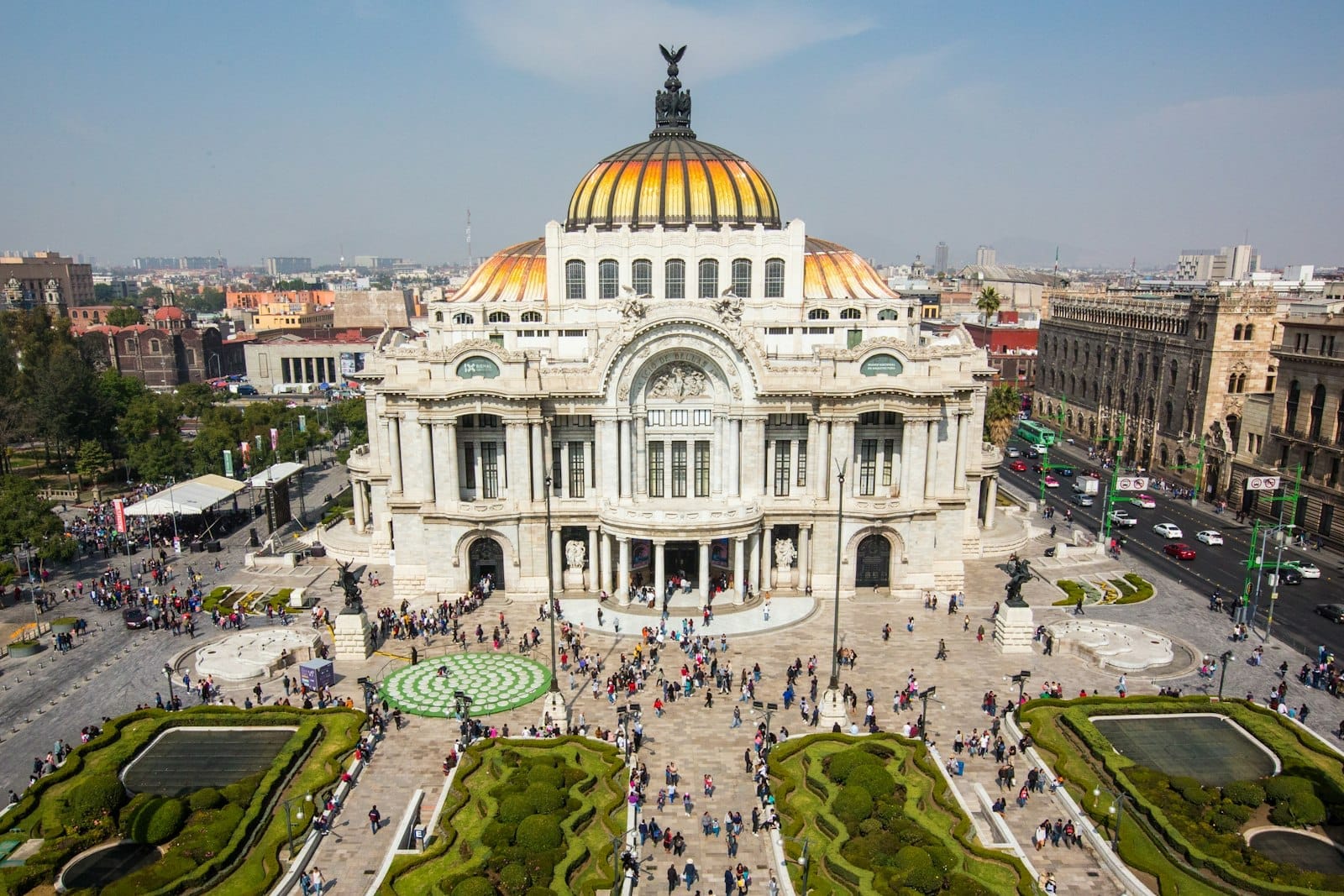 Vista aérea diurna e ensolarada do Palacio de Bellas Artes na Cidade do México, com pessoas caminhando e jardins paisagísticos na frente.
