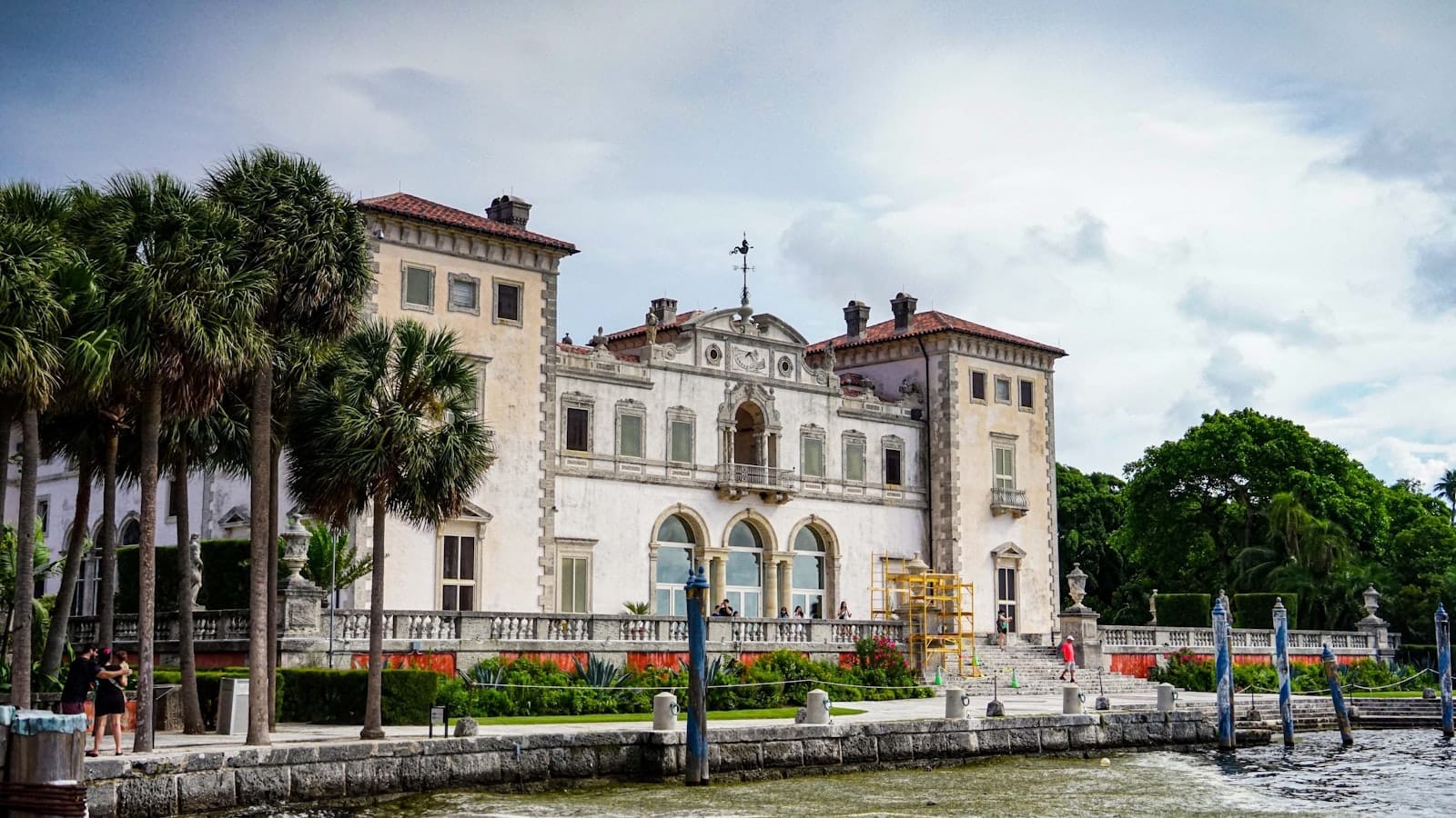 Mansão histórica do Museu e Jardins Vizcaya em Miami, com palmeiras e visitantes ao longo da orla sob um céu nublado.
