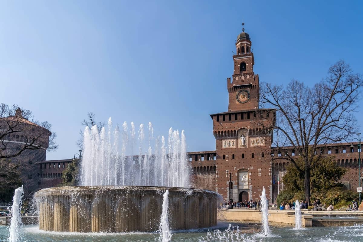 Blick auf das Castello Sforzesco in Mailand mit dem zentralen Uhrenturm und einem großen Brunnen im Vordergrund an einem sonnigen Tag