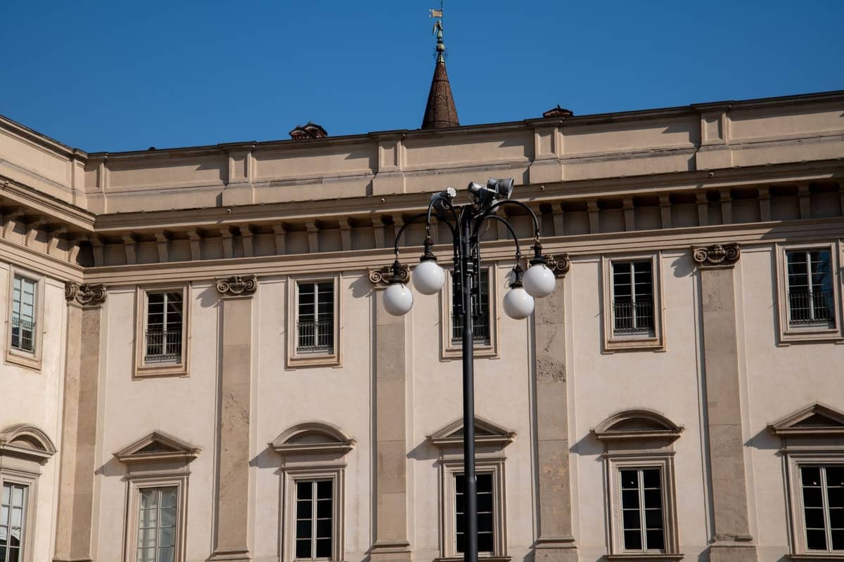 Elegante neoklassizistische Fassade eines historischen Gebäudes in Mailand mit großen Fenstern, Säulen und einer zentralen Straßenlaterne vor blauem Himmel.