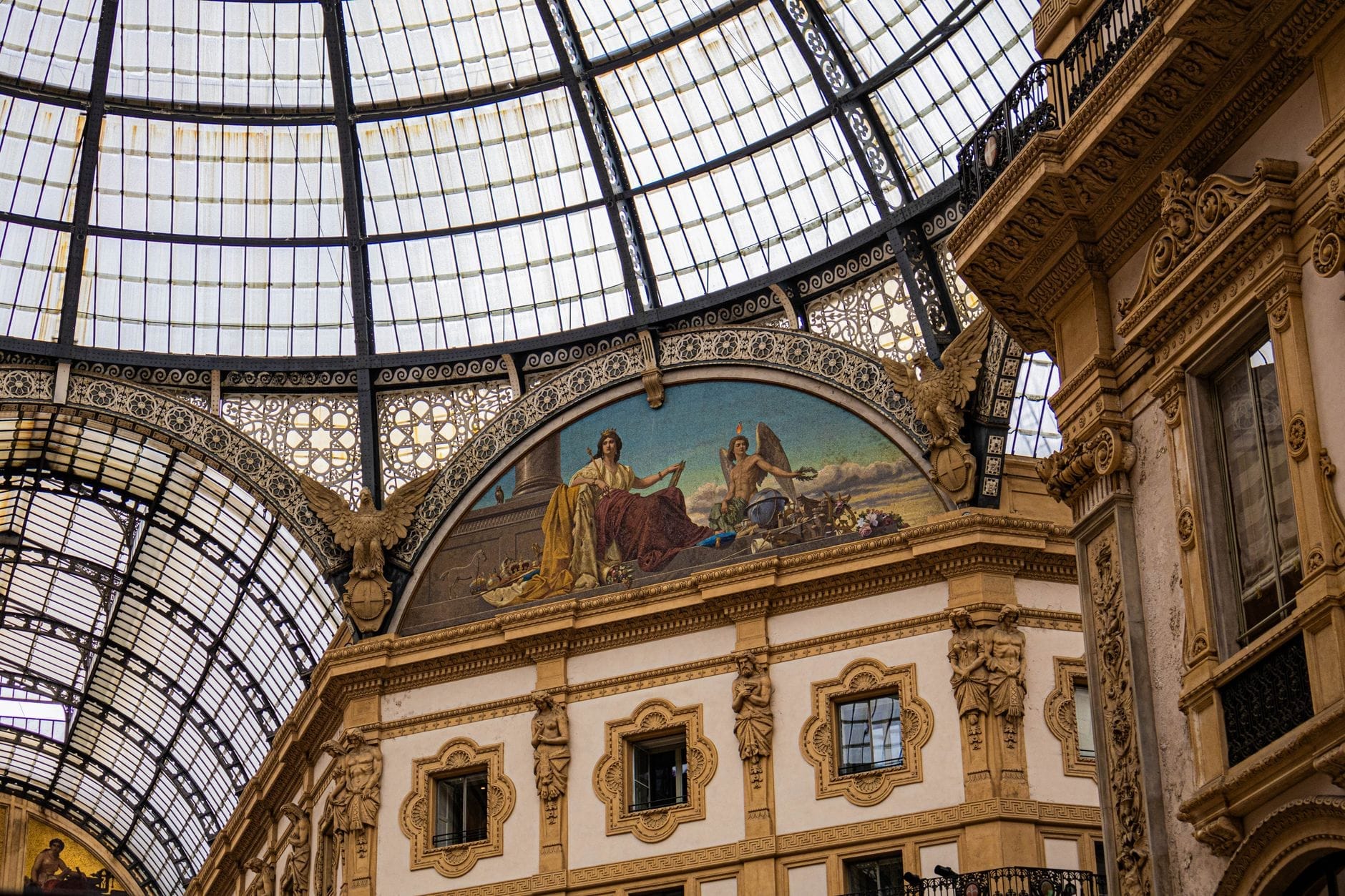 Teto com cúpula de vidro e arquitetura ornamentada no interior da Galleria Vittorio Emanuele II em Milão, com obras de arte clássicas e elementos decorativos detalhados.