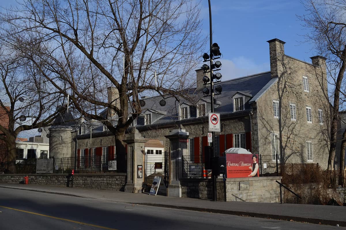 Vista amplia del Museo Château Ramezay en Montreal, un histórico edificio de piedra con contraventanas rojas y un letrero de entrada, visto desde el otro lado de la calle.