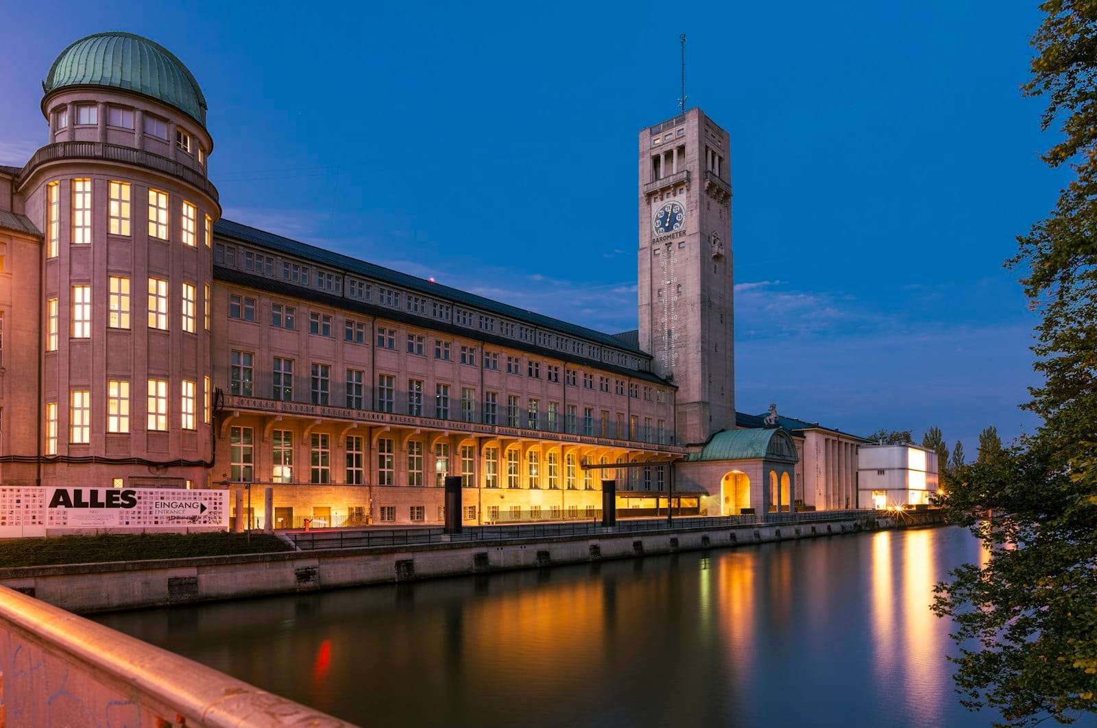 วิวยามเย็นของอาคารประวัติศาสตร์ Deutsches Museum ในมิวนิกที่สะท้อนในแม่น้ำ Isar พร้อมหอนาฬิกาที่ส่องสว่าง
