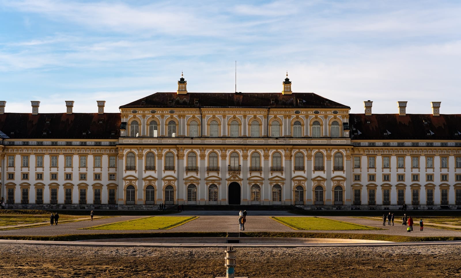 วิวกว้างของด้านนอกสไตล์บาโรกอันยิ่งใหญ่ของพระราชวัง Nymphenburg พร้อมผู้คนเดินอยู่ด้านหน้าในวันที่แดดสดใสที่เมือง Munich