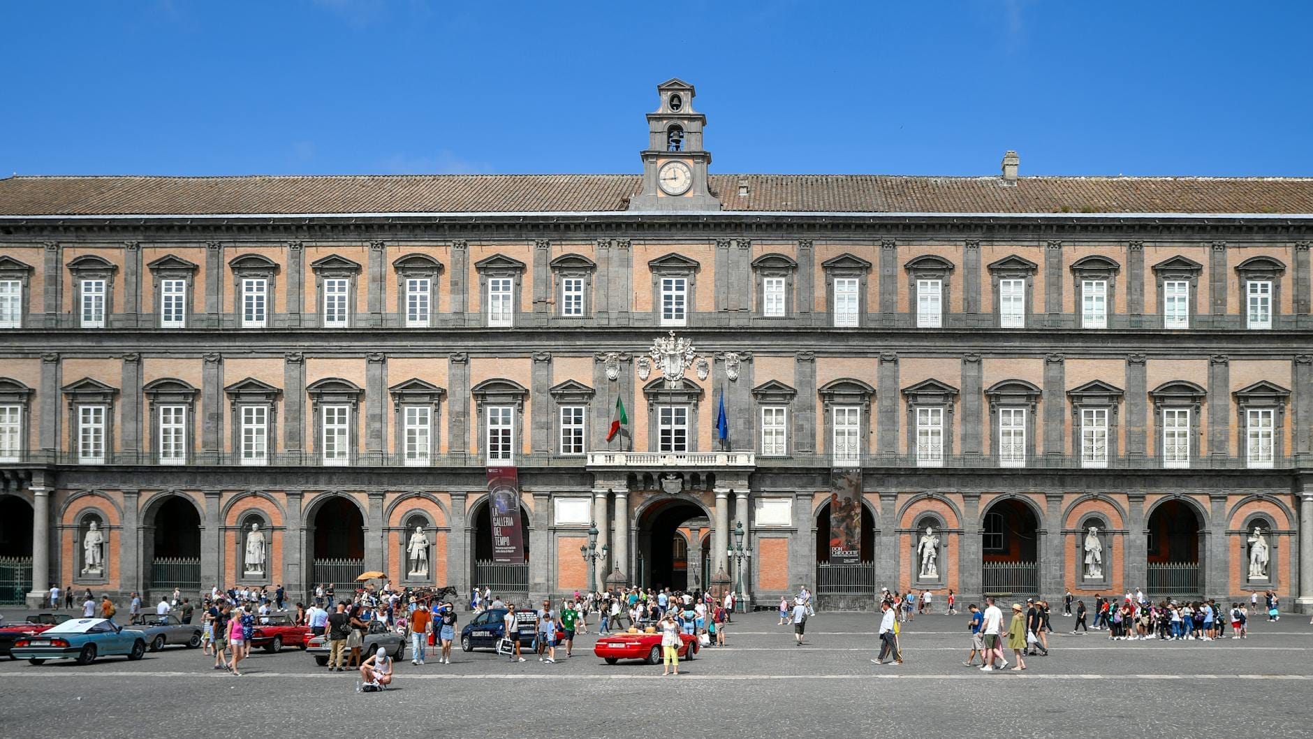 Vista ampla da grande fachada do Palazzo Reale di Napoli com pessoas e carros na praça em frente.