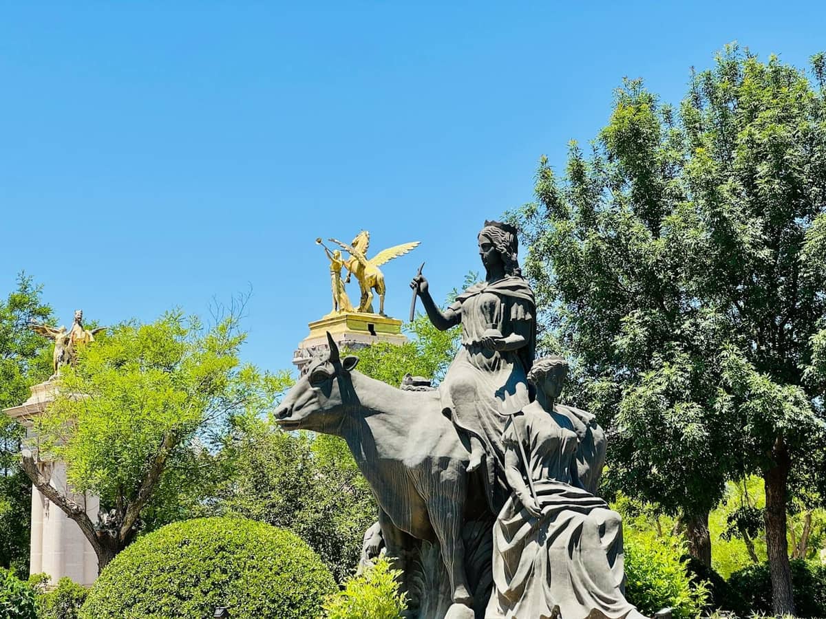 Outdoor sculpture of a seated woman with a bull, surrounded by trees and bushes under a clear blue sky.