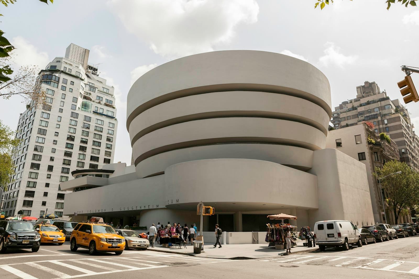 Vista externa do Museu Guggenheim em Nova York, com táxis amarelos e pedestres na movimentada esquina em frente ao icônico edifício moderno.