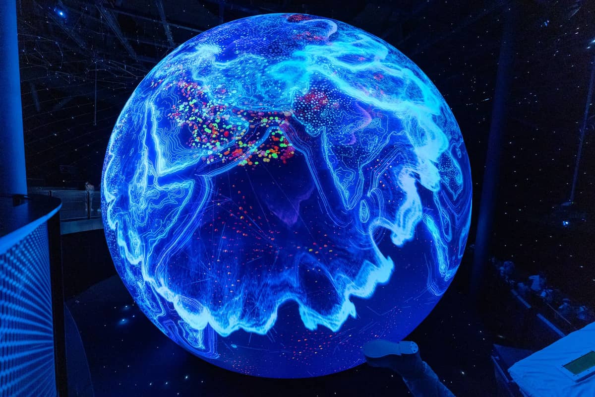Large illuminated digital globe display glowing blue inside a dark science museum with visitors nearby.