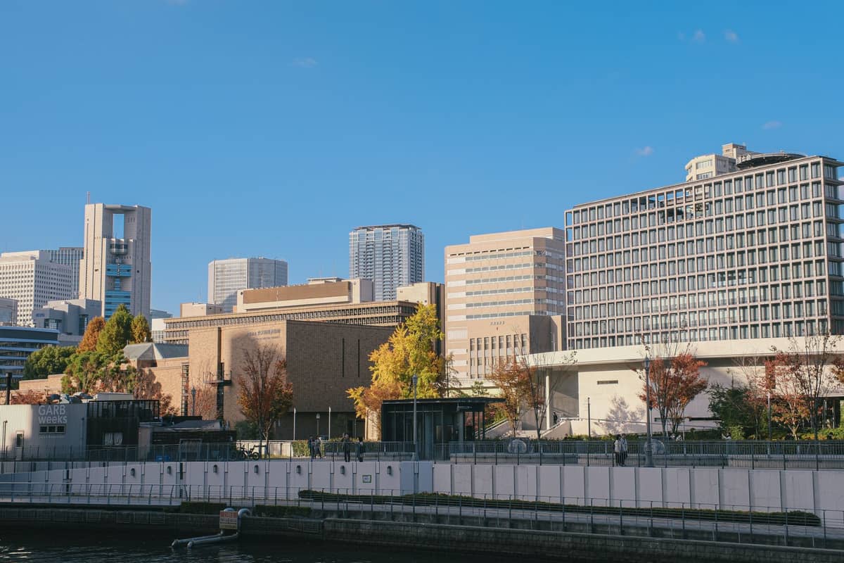 Modern buildings of Nakanoshima seen across the river, including the distinctive Osaka Science Museum and NMAO architecture, under a clear blue sky.
