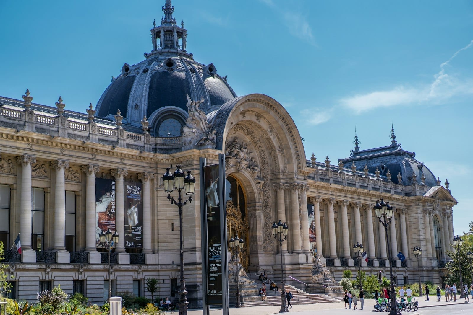 Weitwinkelansicht des prunkvollen Petit Palais Museums in Paris an einem sonnigen Tag, mit Menschen in der Nähe des Eingangs und Bannern an der Fassade.