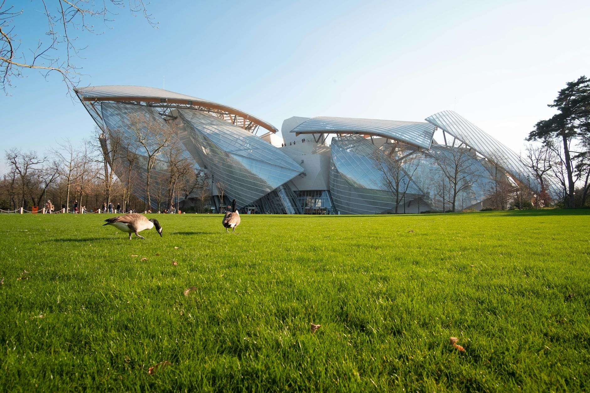 Weitwinkelansicht der beeindruckenden Glas- und Stahlarchitektur des Museums Fondation Louis Vuitton, von einer Rasenfläche mit Bäumen und blauem Himmel aus gesehen.