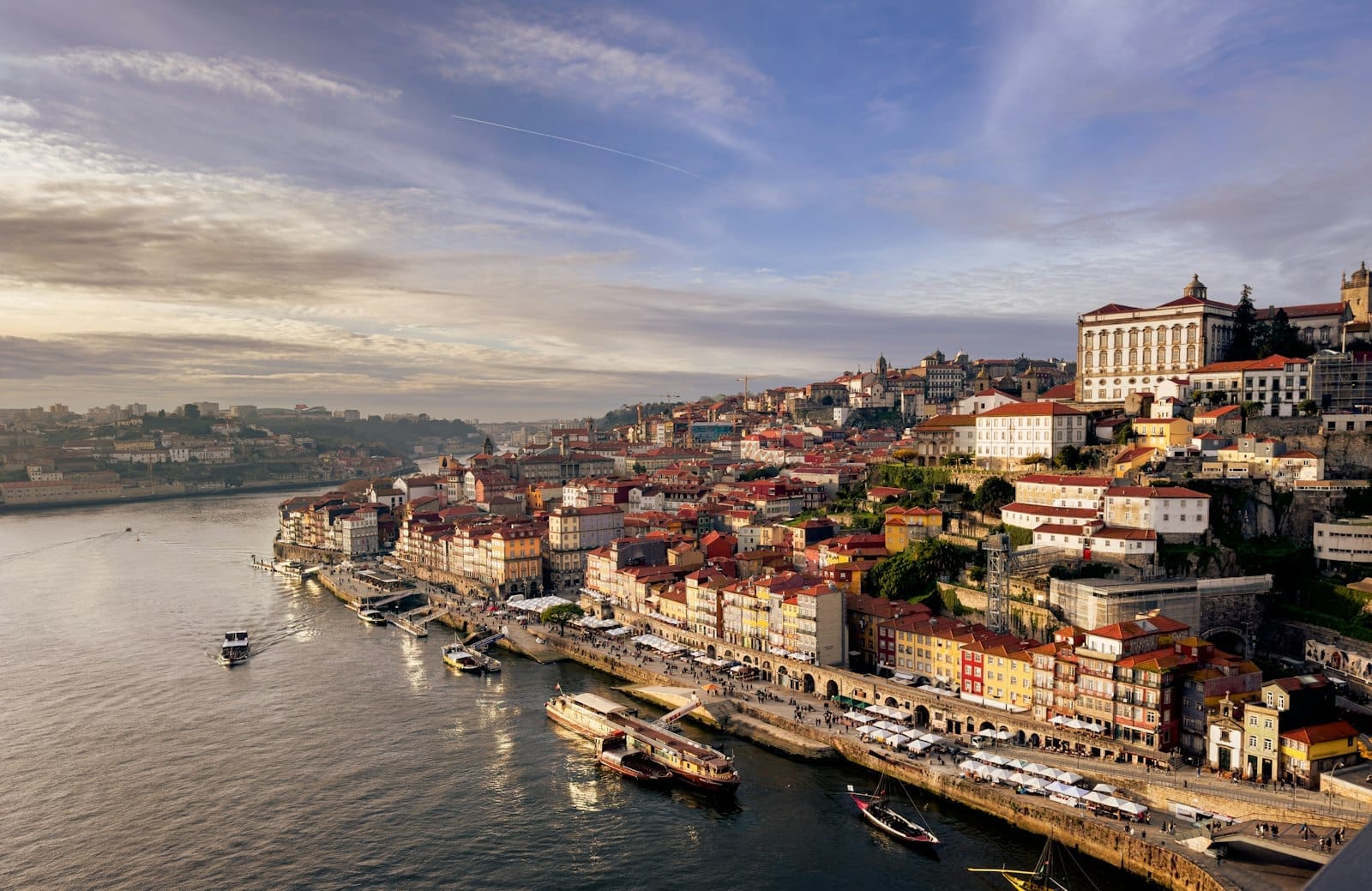 Vista panorâmica das coloridas construções à beira-rio do Porto, com barcos no Rio Douro e os marcos da cidade sob um céu dramático no início da noite.