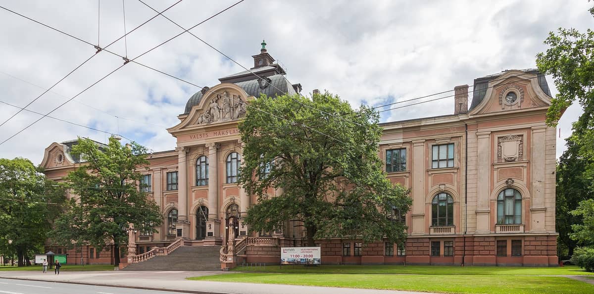 Frontansicht des Lettischen Nationalen Kunstmuseums in Riga – ein imposantes historisches Gebäude mit aufwendigen Architekturdetails, Bäumen und Besuchern davor.