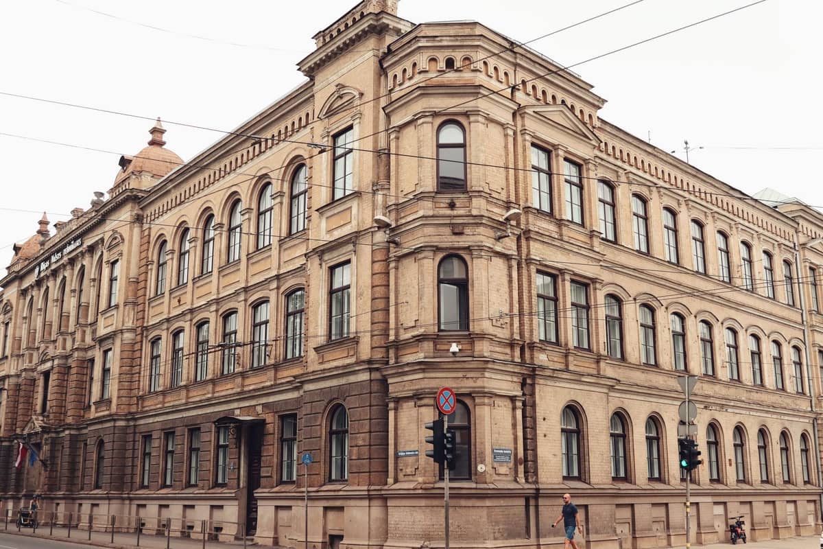 Großes historisches Steingebäude mit Bogenfenstern an einer Ecke in Riga, an einem bewölkten Tag mit Straßenansicht aufgenommen