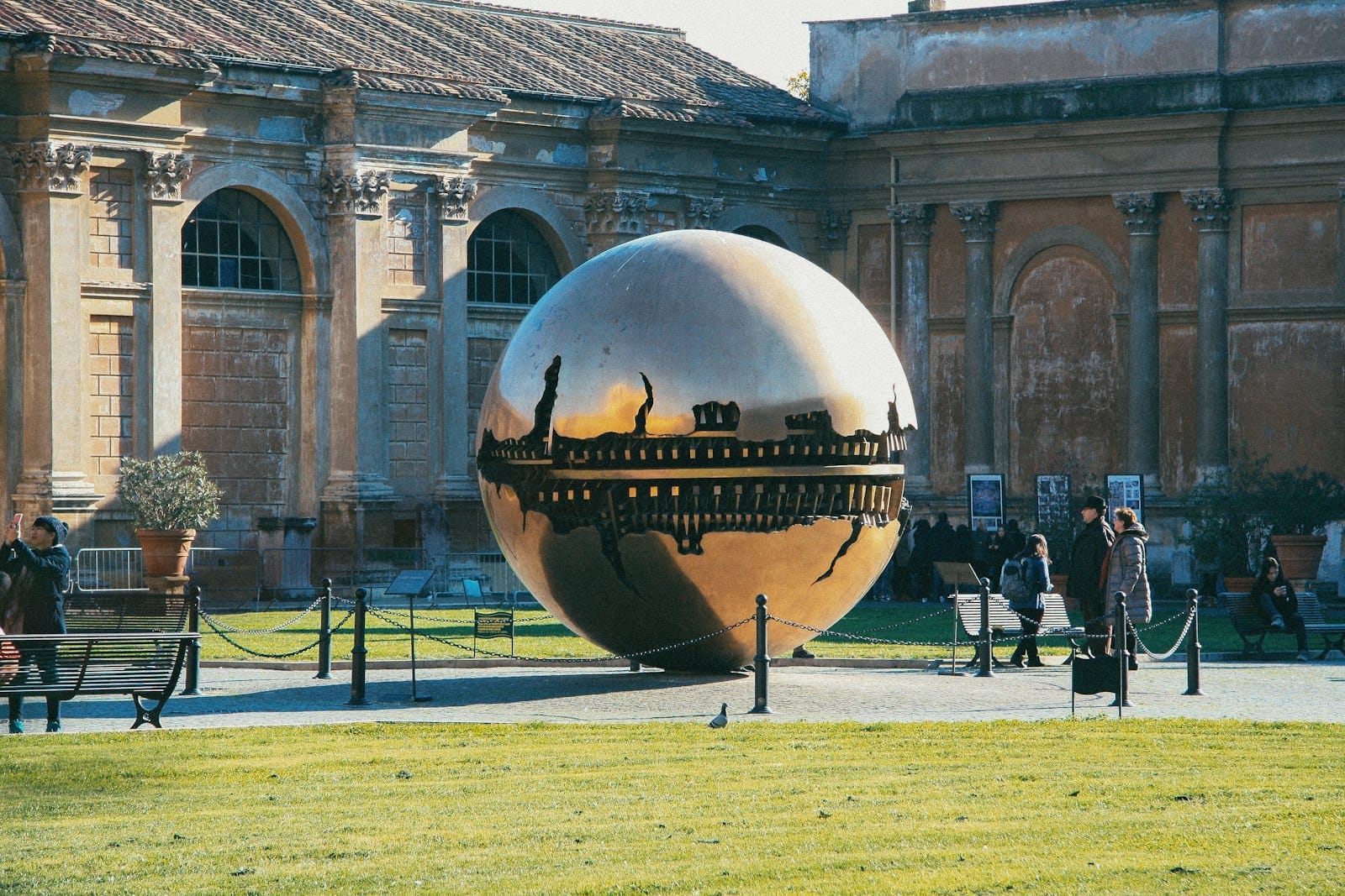 Modern bronze sphere sculpture (Sphere Within a Sphere) in the courtyard of a contemporary museum in Rome, with people walking nearby.