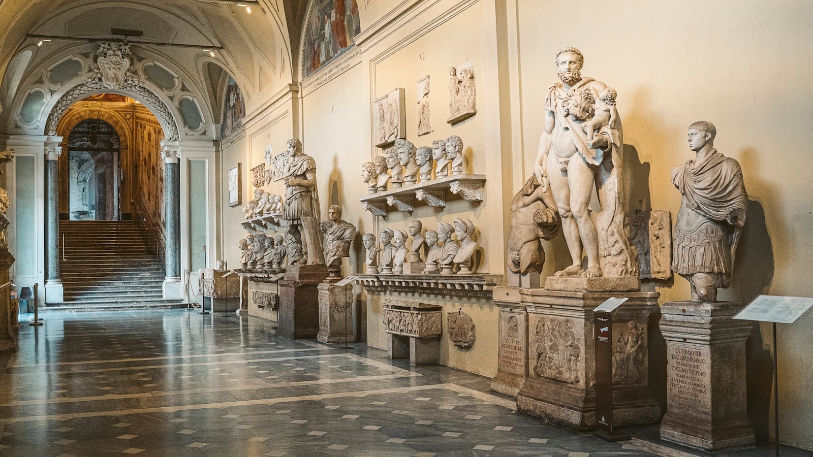 Wide view of a grand museum hall in Rome featuring Roman statues and busts displayed along the walls beneath vaulted ceilings, evoking art and history.
