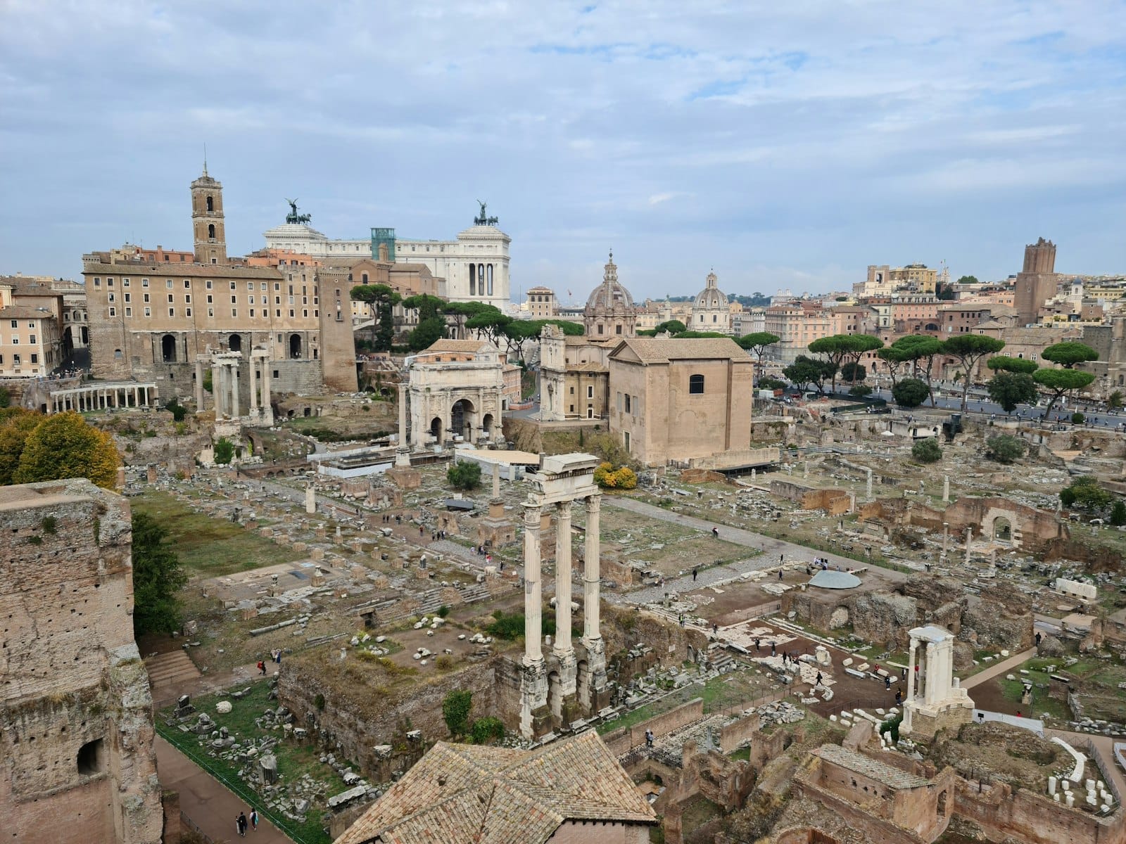 Wide view of the Roman Forum with scattered ancient ruins, columns, and historic buildings under a blue sky in Rome.
