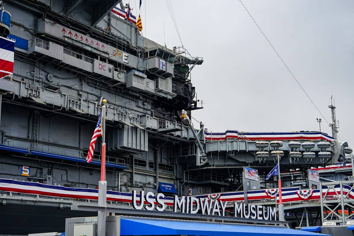 ภาพระยะใกล้ของทางเข้า USS Midway Museum พร้อมธงและโครงสร้างสีเทาขนาดใหญ่ของเรือที่ประดับด้วยธงประดับรักชาติ