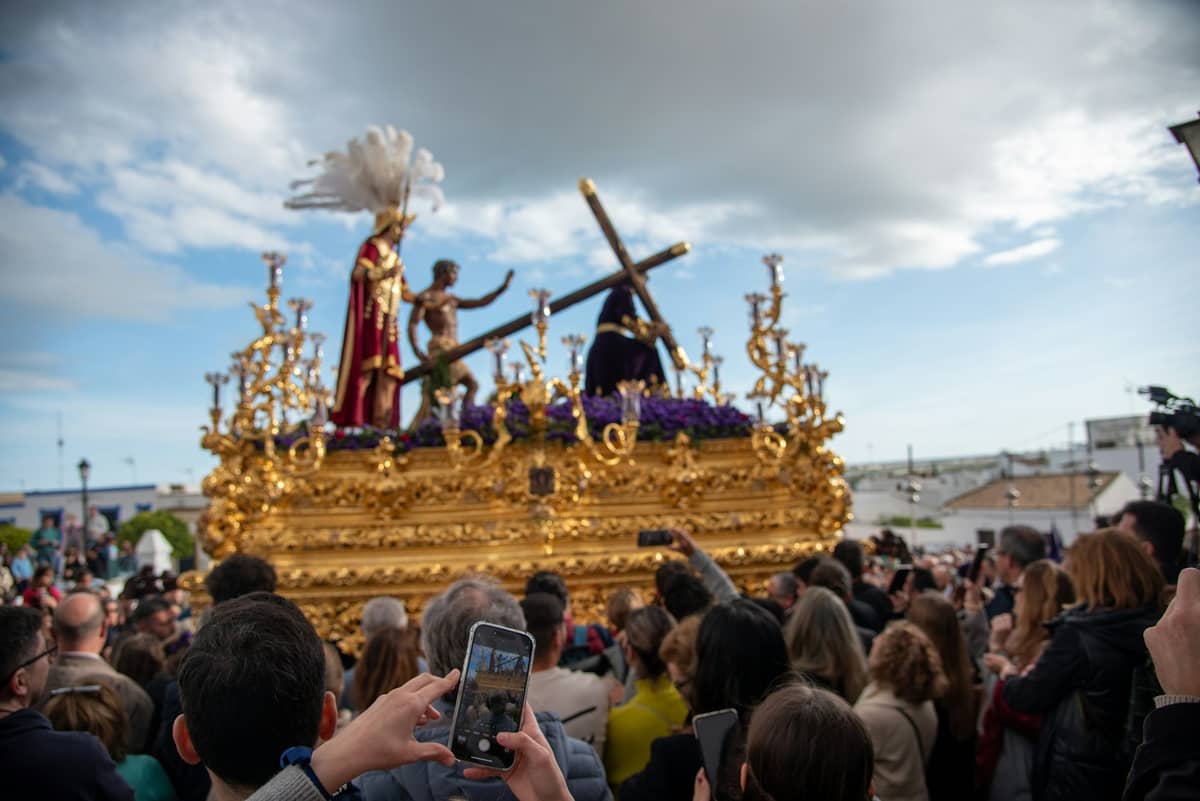 รถแห่ประดับตกแต่งอย่างวิจิตรของ Semana Santa พร้อมการตกแต่งสีทองและรูปปั้นทางศาสนา ล้อมรอบด้วยฝูงชนที่ถ่ายภาพกลางแจ้งใต้ท้องฟ้าครึ้ม