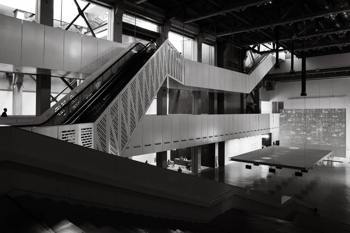 Photo en noir et blanc de l'intérieur d'un musée moderne avec des escalators géométriques, des éléments industriels et un grand espace d'exposition ouvert, évoquant un lieu d'art contemporain.